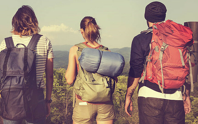 Drei junge Menschen stehen mit Wandergepäck auf einer Anhöhe und sehen in die Ferne. 