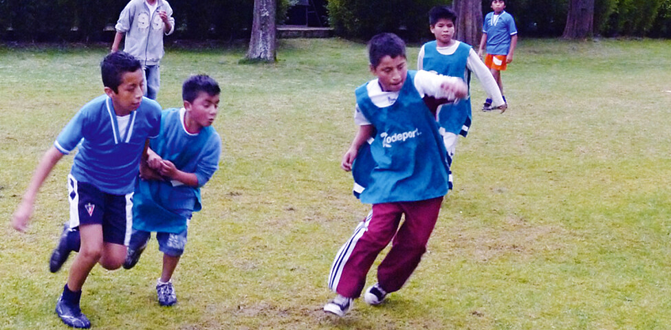 Eine Gruppe junger Buben, die beim Fußballspielen sind. Ein Junge will gerade den Ball schießen, die anderen laufen ihm hinterher.