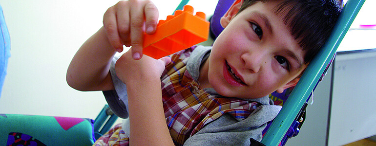Ein junger Bub mit schwarzem Haar sitzt in einem türkisfarbenen Kinderwagen. Er zeigt dabei sein Spielzeug - ein oranger Legobauklotz. 