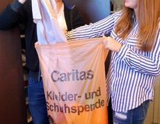 Zwei junge Menschen sind gerade dabei, Kleidung in den orangen Kleidersack zu geben. Sie stehen vor einem braunen Kleiderschrank und lachen.