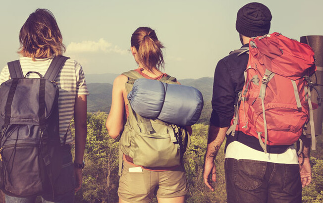 Drei junge Menschen stehen mit Wandergepäck auf einer Anhöhe und sehen in die Ferne. 
