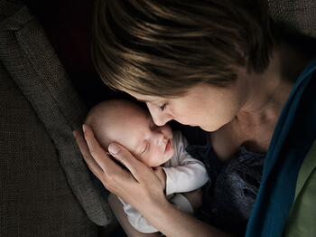 Eine junge Mutter hält ein neugeborenes in ihren Händen. Behutsam liegt der Kopf des Babys in einer ihren Händen. Beide haben die Augen geschlossen und es scheint, als genießen sie die Situation.
