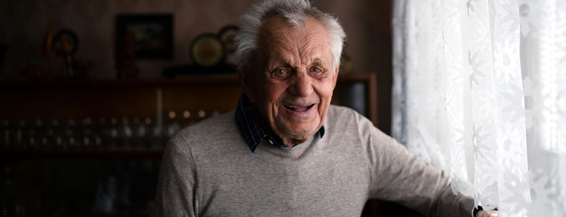 Ein lachender Mann im hohen Alter steht an einem Licht durchfluteten Fenster.
