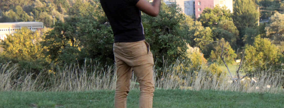 Ein junger Flüchtling steht mit dem Rücken zur Kamera in einer Wiese. Er hält sein Handy in die Luft und es scheint, als würde er ein Foto machen. Er trägt eine beige Hose, ein schwarzes T-shirt und eine Kappe.
