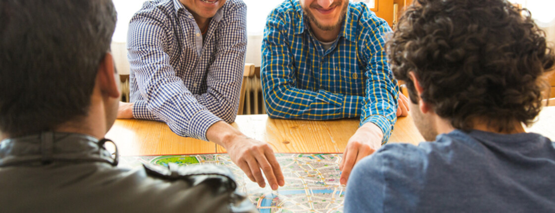 Unbegleitet minderjährige Flüchtlinge spielen gemeinsam ein Brettspiel. Sie sitzen in einem Freizeitraum, der gut ausgeleuchtet ist, an einem Holztisch. Die vier Männer sind dabei sichtlich erfreut und genießen es.