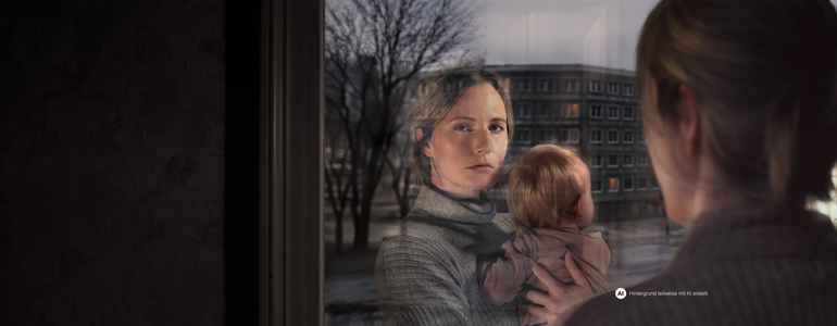 Eine Frau mit Kind am Arm schaut aus dem Fenster. Ihr Gesicht spiegelt sich im Fensterglas wider. Vor dem Fenster ist eine Wohngegend erkennbar.