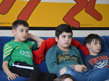 Drei Kinder sitzen auf dem Sofa und hören einer Vorleserin zu.