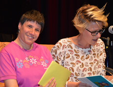 Eine Frau mittleren Alters liest etwas aus einem Buch vor. Neben ihr sitzt eine Frau mittleren Alters mit Beeinträchtigung. 