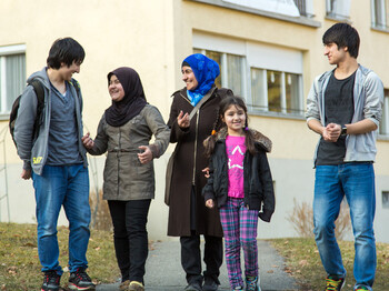 Eine Flüchtlingsfamilie befindet sich vor ihrem Quartier, in dem sie untergebracht sind. Es ist in diesem Fall Gaisbühel in Schlins bzw. in Bludesch. Die Mutter unterhaltet sich mit ihren vier Kindern. Jeweils zwei Töchter und Söhne. Sie sind alle schlicht gekleidet und tragen einfärbige Jacken und t-Shirts. Zusätzlich tragen die Mutter und ihre älteste Tochter ein Kopftuch. Die Stimmung ist angenehm und wohlwollend. Alle lachen einander an und sind fröhlich.