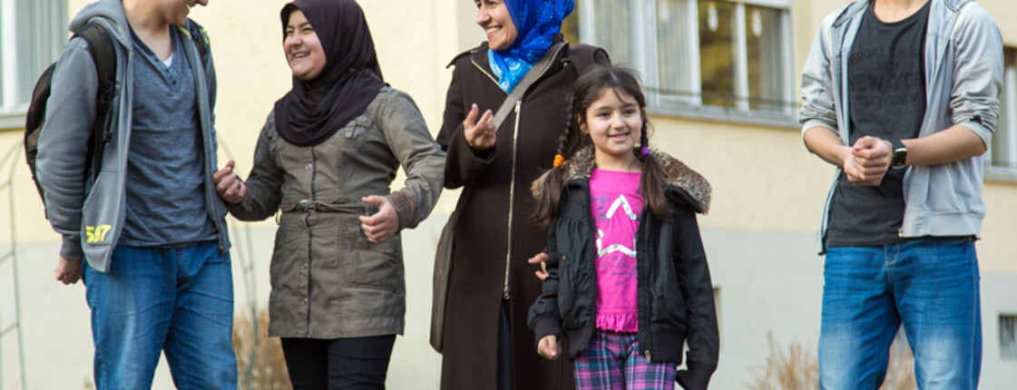 Eine Flüchtlingsfamilie befindet sich vor ihrem Quartier, in dem sie untergebracht sind. Es ist in diesem Fall Gaisbühel in Schlins bzw. in Bludesch. Die Mutter unterhaltet sich mit ihren vier Kindern. Jeweils zwei Töchter und Söhne. Sie sind alle schlicht gekleidet und tragen einfärbige Jacken und t-Shirts. Zusätzlich tragen die Mutter und ihre älteste Tochter ein Kopftuch. Die Stimmung ist angenehm und wohlwollend. Alle lachen einander an und sind fröhlich. 