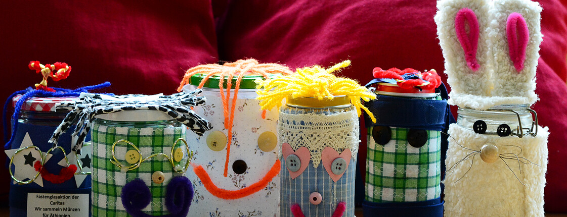 Auf einem Tisch stehen 5 Marmeladengläser, welche von Kindern der Volksschule Dornbirn Haselstauden gestaltet wurden. Die Gläser wurden mit verschiedenen Stoffen und Knöpfen auch mit einem Gesicht bestückt. Ein Kind bastelte aus dem Glas einen Hasen.