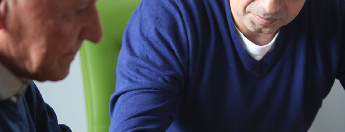 Mann in blauem Pullover sitzt mit älterem Herren an einem Tisch und erklärt etwas.
