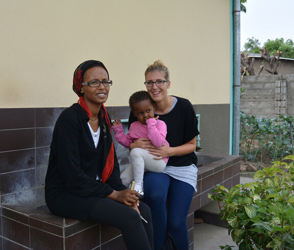 Caritas-Mitarbeiterin Nicole Heim zu Besuch im Girls Hostel in Äthiopien. Sie sitzt mit einer Frau und einem Kind auf dem Schoß auf der Kante eines Brunnens im Freien. Alle bis auf das junge Mädchen sind dunkel bzw. schwarz gekleidet. Das Kind trägt ein rosarotes Oberteil mit einer beigen Strumpfhose. 