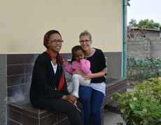 Caritas-Mitarbeiterin Nicole Heim zu Besuch im Girls Hostel in Äthiopien. Sie sitzt mit einer Frau und einem Kind auf dem Schoß auf der Kante eines Brunnens im Freien. Alle bis auf das junge Mädchen sind dunkel bzw. schwarz gekleidet. Das Kind trägt ein rosarotes Oberteil mit einer beigen Strumpfhose. 