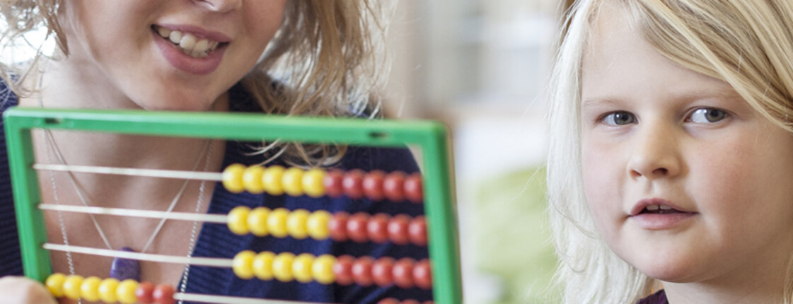 Eine junge Frau, die freiwillig bei der Caritas mitarbeitet, sitzt mit einem kleinen blonden Mädchen an einem Tisch und erklärt ihm anhand eines Rechenhilfsmittel, dem Abakus, das Zählen.