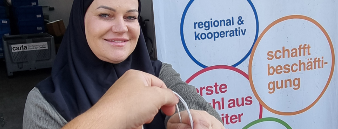 Eine Frau hält eine Tragetasche mit gespendeter Kleidung und steht neben einem Plakat der Sachspendenannahme von Carla mit den Sprüchen "Regional und Kooperativ" und "Carla schafft Beschäftigung".