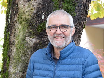 Lächelnder älterer Freiwilliger der Caritas in blauer Jacke vor Baum.