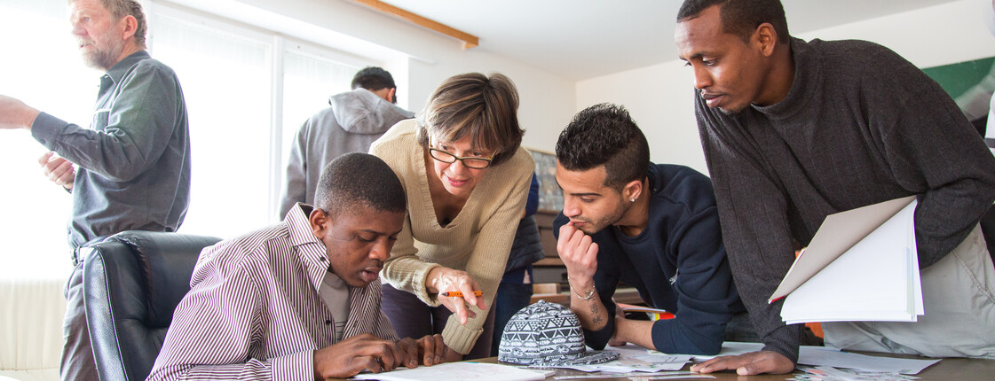 Bildungsangebote für Flüchtlinge Flüchtlinge aus Afrika und dem nahen Osten lernen Deutsch. Sie sitzen gespannt und konzentriert an einem Tisch und blicken auf ihre Unterlagen. Eine Lehrerin hilft ihnen dabei den Stoff besser aufzunehmen und steht für jede Frage offen.