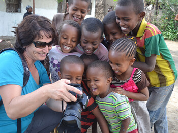Caritas-Mitarbeiterin Elke Kager zeigt äthiopischen Kindern Fotos auf ihrer Kamera. Die Kinder schauen gespannt und sind erfreut. Elke trägt eine blaue Bluse und die Kinder sind bunt, in unterschiedlichen Farben gekleidet. 