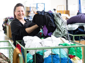 Foto Mathis Eine carla Mitarbeiterin mittleren Alters mit langen schwarzen Haaren sortiert fröhlich und heiter Kleidung aus einem grünen Schiebewagen.