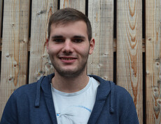 Ein junger Mann steht vor einem Holzhintergrund. Er lächelt in die Kamera und trägt ein weißes T-shirt mit einer blauen Kaputzenjacke darüber.