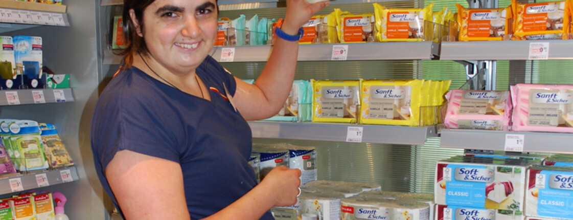 Bianca Panek, eine junge erwachsene Frau mit schwarzen langen Haaren und einem dunkelblauen t-Shirt, ist gerade am Einräumen von Waaren in einem Drogeriemarkt.