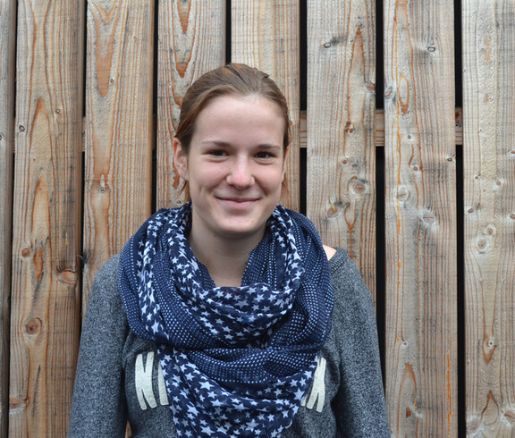 Eine junge Dame steht vor einem Holzhintergrund für ein Portraitfoto. Sie trägt ein blaues Halstuch und ihre Haare zu einem Pferdeschwanz zusammen. 