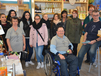 Menschen mit und ohne Behinderung stehen zu einem Gruppenfoto zusammen. Ein Mann sitzt in einem Rollstuhl. Sowohl junge als auch ältere Personen sind vertreten. Sie sind in einem Einkaufsladen. 