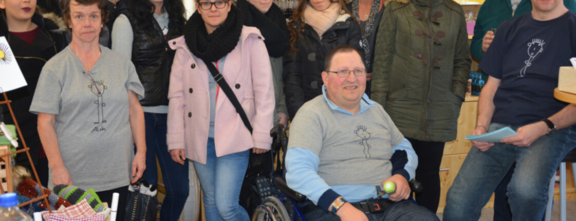 Menschen mit und ohne Behinderung stehen zu einem Gruppenfoto zusammen. Ein Mann sitzt in einem Rollstuhl. Sowohl junge als auch ältere Personen sind vertreten. Sie sind in einem Einkaufsladen. 