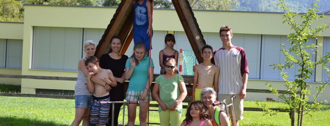 Kinder und Jugendliche stehen im Freien für ein Gruppenfoto zusammen. Sie befinden sich in einer Wiese, vor einem kleinen Holzhaus. 