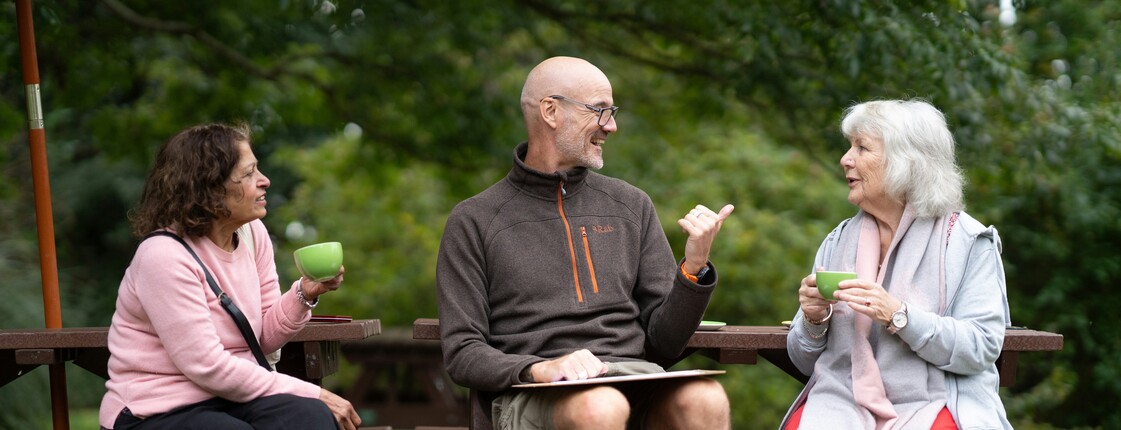 Zwei gut gelaunte Seniorinnen und ein junger Mann sitzen auf Parkbänken und unterhalten sich.