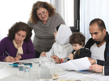Eine Frau mittleren Alters lernt mit einer Flüchtlingsfamilie. Auf dem Tisch befinden sich Blätter, Bücher und Stifte. Die Frau steht dabei hinter der Familie. Die Familie sitzt am Tisch.