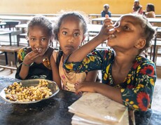 Drei Mädchen sitzen an einem Tisch in einer Schulkantine und essen gemeinsam Mais und Bohnen.