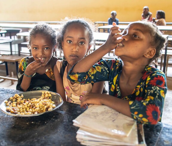 Drei Mädchen sitzen an einem Tisch in einer Schulkantine und essen gemeinsam Mais und Bohnen.