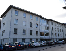 An einer Straße steht ein dreistöckiges größeres Gebäude, das weder alt noch neu in seiner Erscheinung ist. Die Aussenfassade ist weiß. Vor dem Block stehen etliche Autos in einer Reihe auf Parkplätzen. 