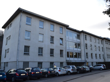 An einer Straße steht ein dreistöckiges größeres Gebäude, das weder alt noch neu in seiner Erscheinung ist. Die Aussenfassade ist weiß. Vor dem Block stehen etliche Autos in einer Reihe auf Parkplätzen.