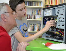Generationenaustausch: Ein junger Bursche mit grauem t-Shirt und ein älterer Mann mit rotem Polo reparieren einen Computer. Die jeweiligen Hardwareteile sind dabei gut ersichtlich. Der PC steht auf einem grünen Tisch. 