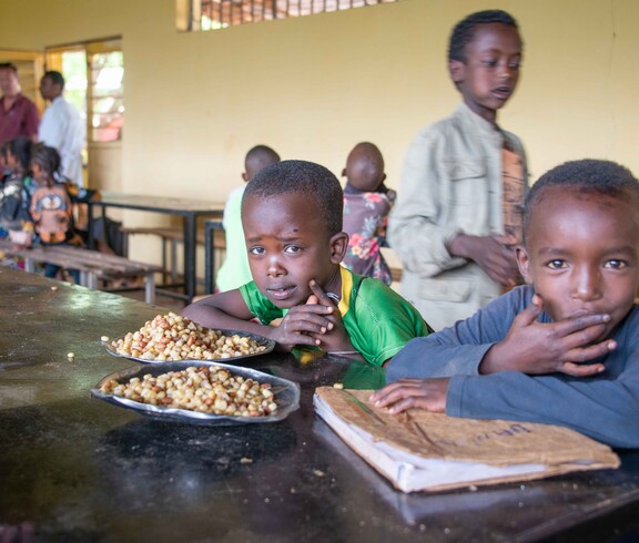 Zwei Kinder sitzen an einem Holztisch in einem Speisesaal und schauen in die Kamera; vor ihnen liegen Teller mit gekochten Maisbohnen.