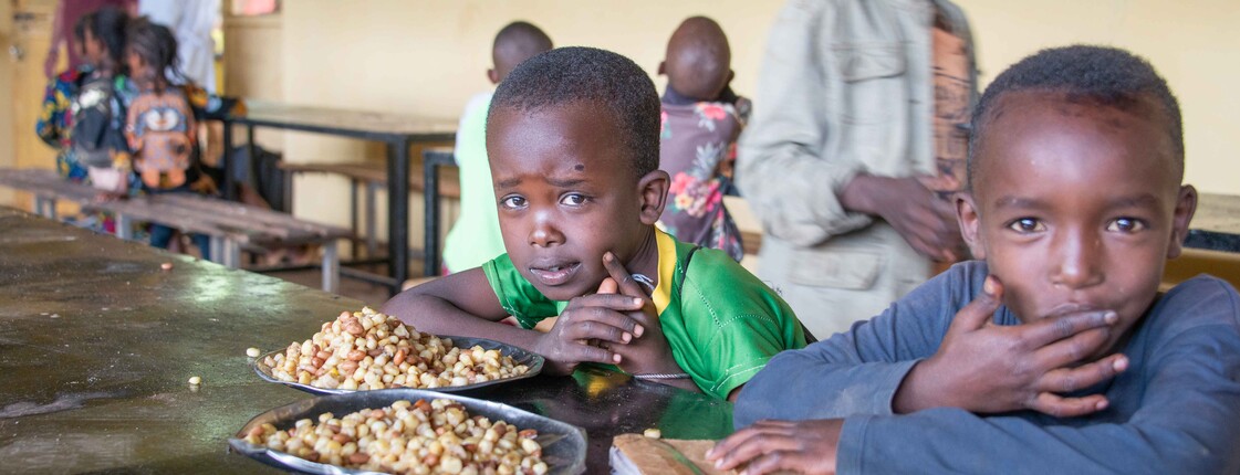 Zwei Kinder sitzen an einem Holztisch in einem Speisesaal und schauen in die Kamera; vor ihnen liegen Teller mit gekochten Maisbohnen.