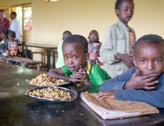 Zwei Kinder sitzen an einem Holztisch in einem Speisesaal und schauen in die Kamera; vor ihnen liegen Teller mit gekochten Maisbohnen.