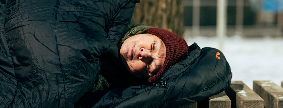 Eine Frau schläft in einen schwarzen Schlafsack gewickelt auf einer Parkbank. Sie hat einen Anorak an und trägt eine rote Haube.