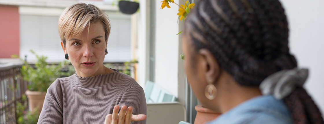 Eine Frau mit kurzen Haaren unterhält sich auf einem Balkon mit einer Frau, die ein Jeanshemd trägt.
