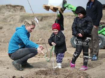 Kinder mit Behinderung der Tagesstätte „Emils Kleine Sonne“ haben gemeinsam mit HelferInnen den Garten des Neubaus bepflanzt Zwei armenische Kinder planzen mit Hilfe einer Freiwilligen einen Baum in der Landschaft. Alle sind dabei in warmen Winterklamotten gehüllt und dunkel bekleidet. Die junge Frewillige trägt im Kontrast zu den anderen drei sich auf dem Foto befindenen Personen einen hellblauen Sweater. Die Kinder sind angesichts der Situation und der Aufmerksamkeit, die sie erfahren, erfreut.