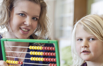 Eine junge Frau, die freiwillig bei der Caritas mitarbeitet, sitzt mit einem kleinen blonden Mädchen an einem Tisch und erklärt ihm anhand eines Rechenhilfsmittel, dem Abakus, das Zählen.