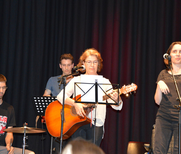Eine Frau mittleren Alters spielt auf einer Gitarre. Im Hintergrund sind noch drei Menschen. Ein junger Mann spielt auf dem Schlagzeug.