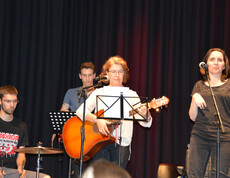 Eine Frau mittleren Alters spielt auf einer Gitarre. Im Hintergrund sind noch drei Menschen. Ein junger Mann spielt auf dem Schlagzeug.
