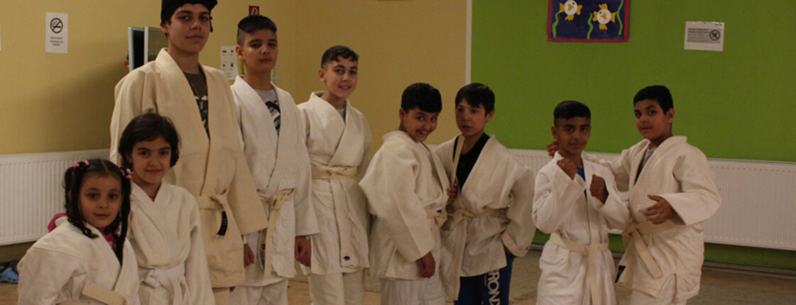 Die Flüchtlingskinder beim Judotraining. Alle tragen eine weiße Judouniform und stehen in einer Reihe. Manche schon in einer Judostellung. 