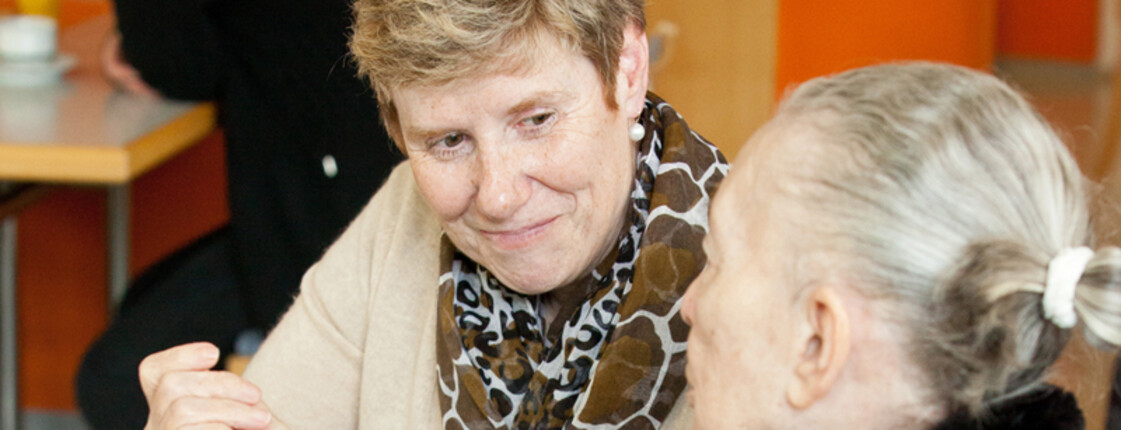Eine Dame mittleren Alters hält ihren Kopf in Richtung einer etwas älteren Dame. Sie grinsen sich gegenseitig an. Die Frau an der linken Seite trägt ein Namensschild.