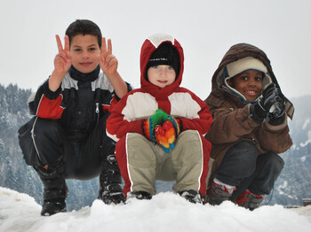 Drei Kinder unterschiedlicher Ethnien spielen im Schnee. Sie tragen dicke Winterbekleidung, die schwarz, weiß, rot und braun ist. Sie sitzen dabei in einer Hocke auf einem Schneehügel. Alle drei Kinder sind erfreut und haben Spaß. Einer der drei Jungs zeigt noch das Victory Zeichen mit beiden Händen. 