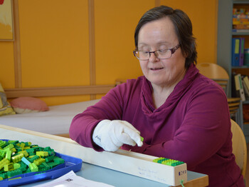 Eine Frau mittleren Alters ordnet Legosteine in eine Holzkiste. Sie trägt weiße Handschuhe und eine Brille.